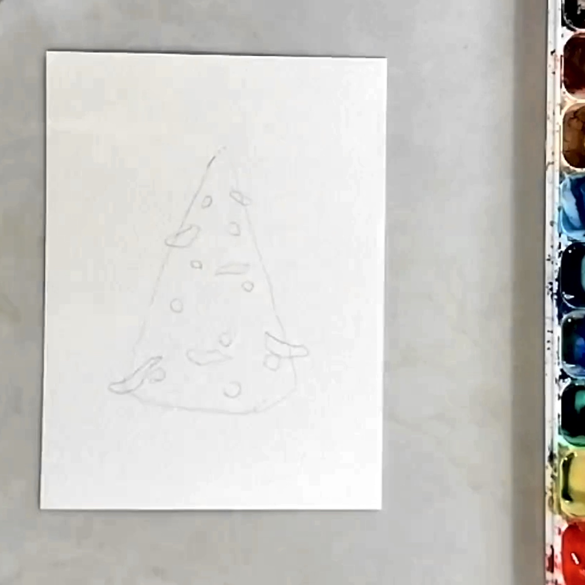 A sketch of a Christmas tree, ready to paint, next to a watercolor palette. 