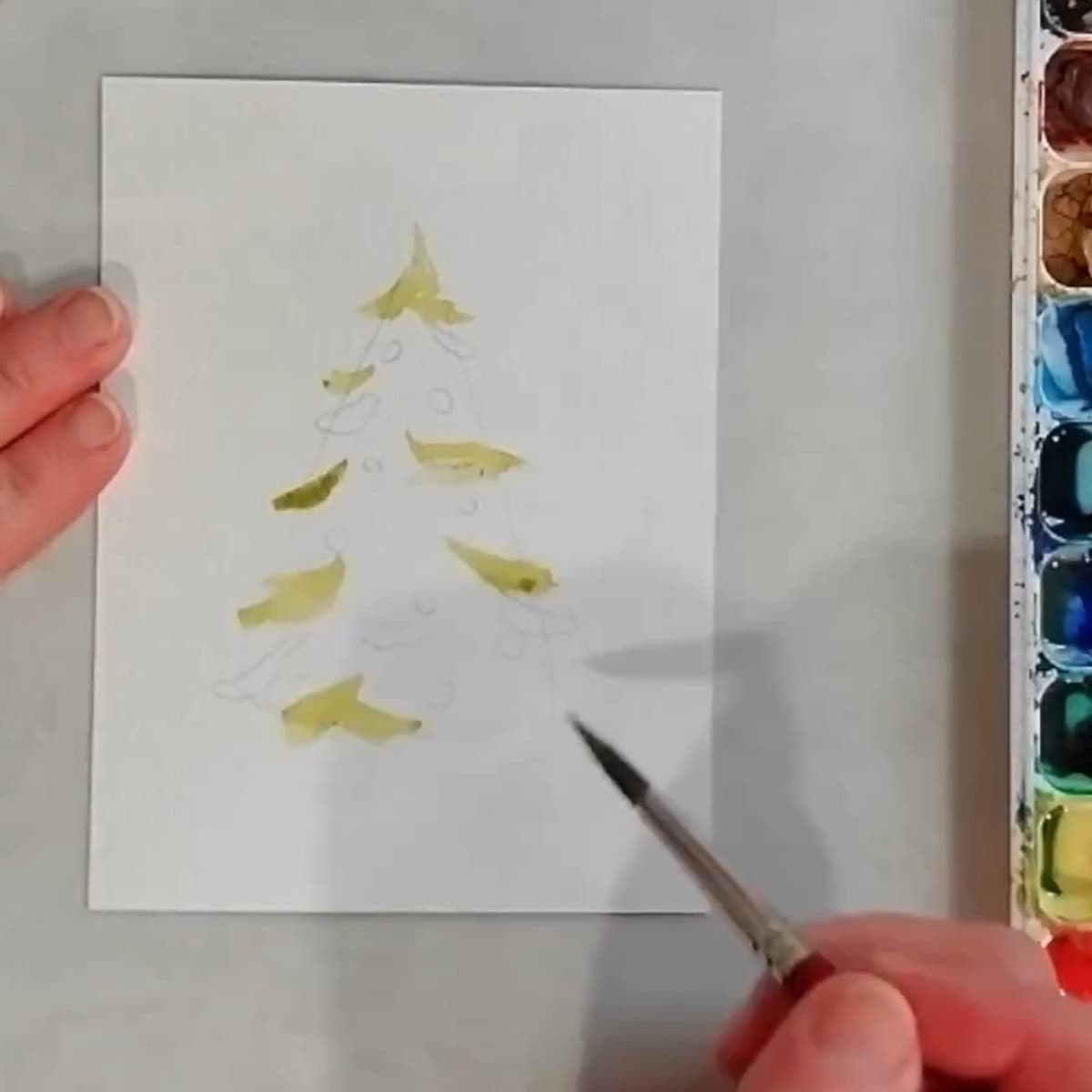 An artist's hand begining to paint a Christmas tree in watercolor.