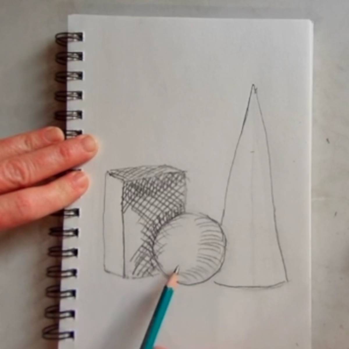 Sketch of a cone, sphere, and box with artist adding cross hatching lines on a spiral ring notebook. 