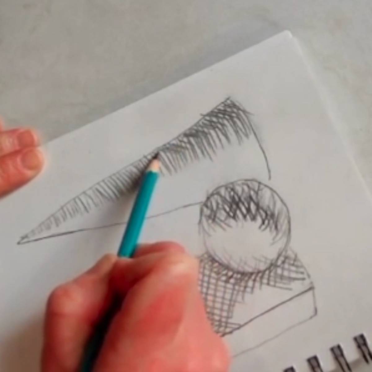 Artist adding cross hatching lines to a sketch of a cone, sphere, and box on a spiral ring notebook.