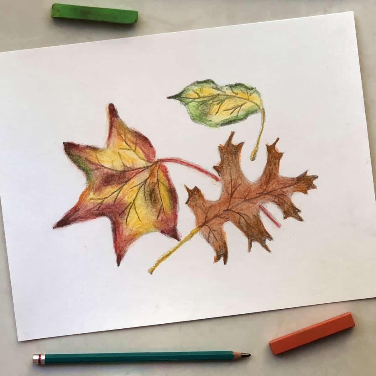Colorful fall leaf drawing created with pencil and soft pastels next to a drawing pencil and orange and green soft pastel sticks.