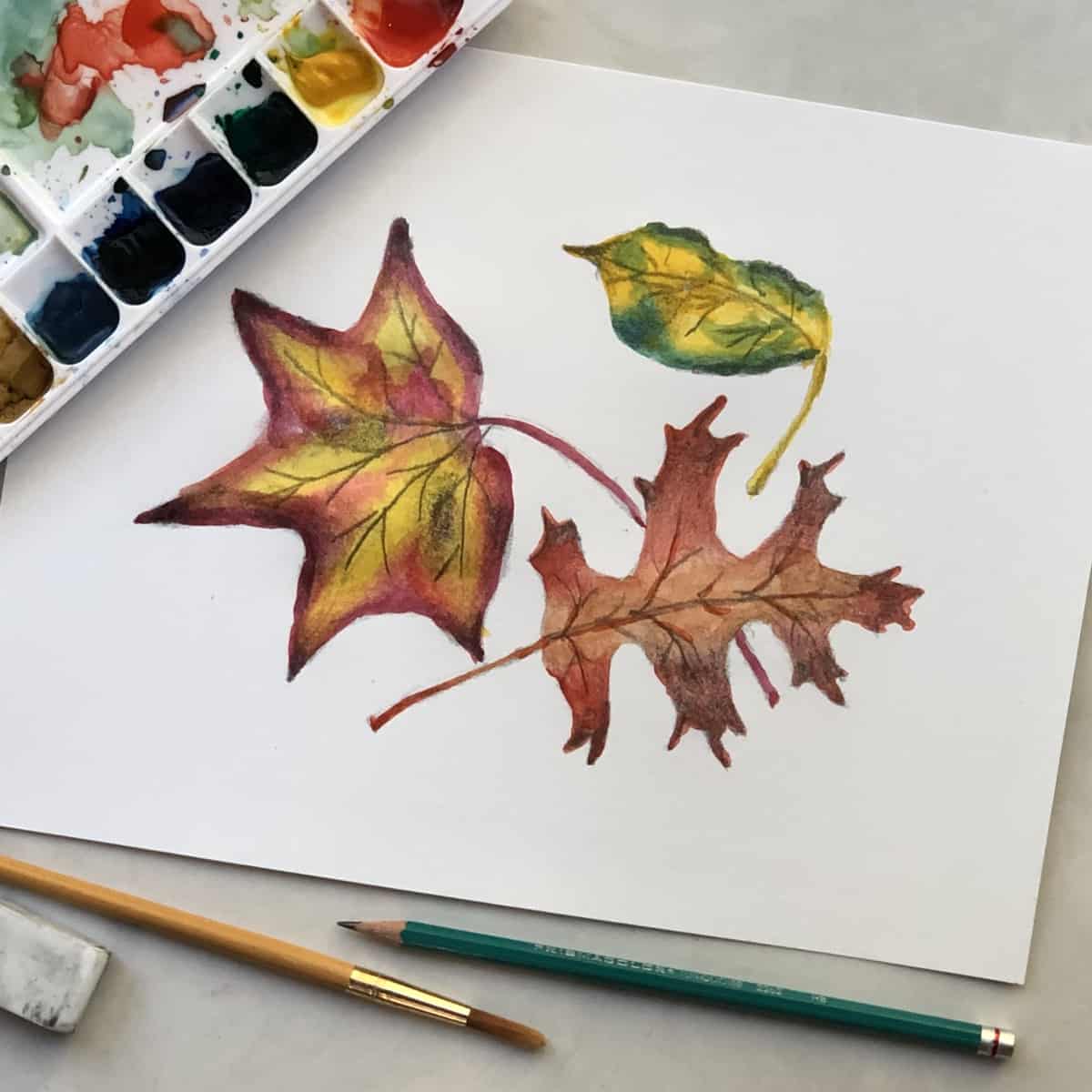 Colorful drawing of fall leaves colored with watercolor paints on paper next to a paintbrush, drawing pencil, eraser, and a watercolor paint set.