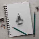 Pencil drawing of a pear on a spiral-bound sketchbook with drawing pencils, pencil sharpener, and sharpening dust.