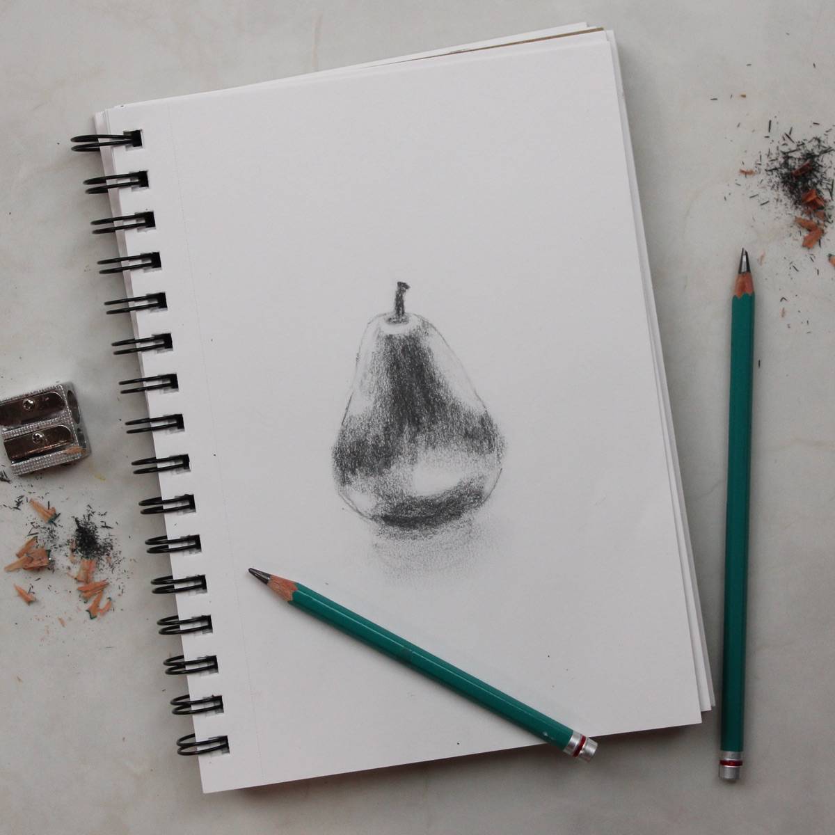 Pencil drawing of a pear on a spiral-bound sketchbook with drawing pencils, pencil sharpener, and sharpening dust. 