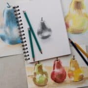 Pencil drawing of a pear on a spiral bound sketchbook next to watercolor painting and colored pencil sketches of pears.