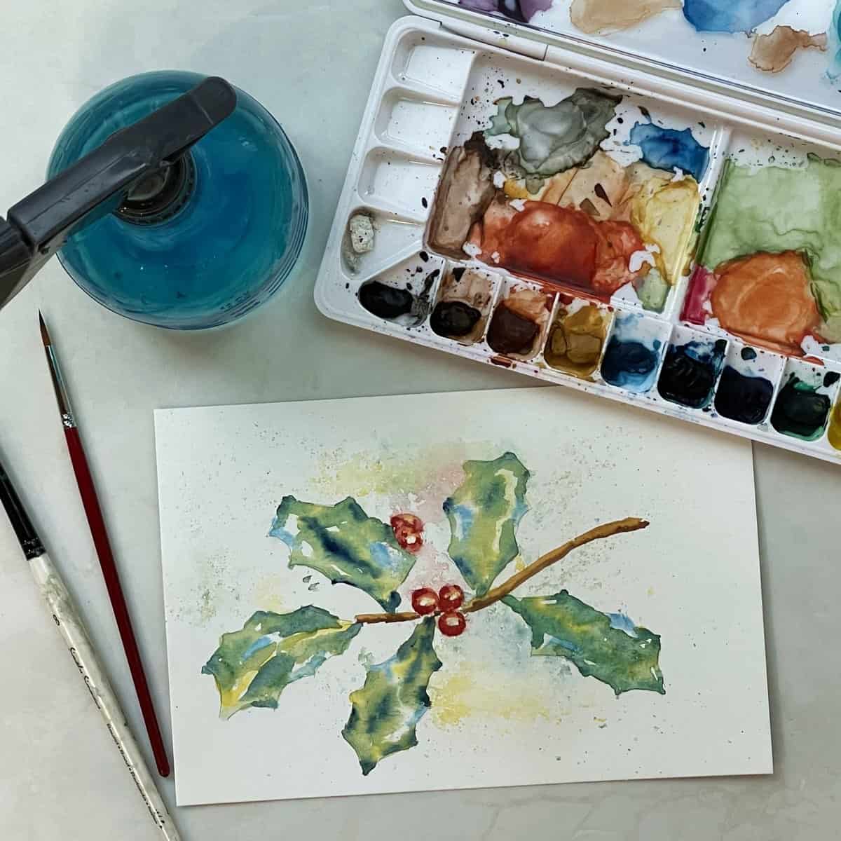 Watercolor painting of holly leaves and berries with splatter effects next to paint brushes, a paint palette and a squirt bottle.