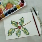 Watercolor painting of a spring of holly with paint pallet and paint brushes.