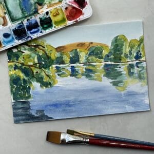 Painting of a watercolor lake scene with trees, a hill and reflections on the water next to paint brushes and a watercolor palette.
