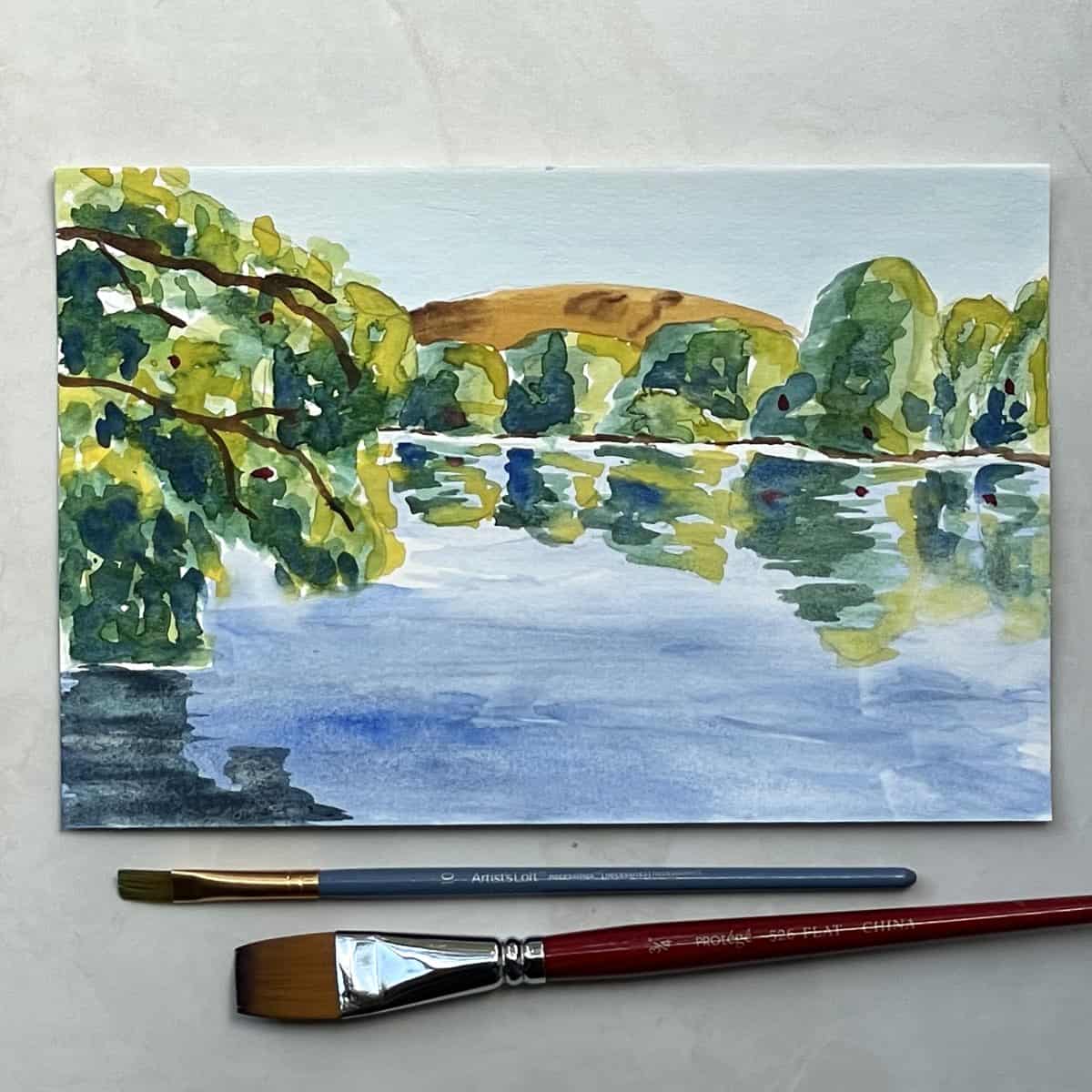 Painting of a watercolor lake scene with trees, a hill and reflections on the water next to paint brushes.