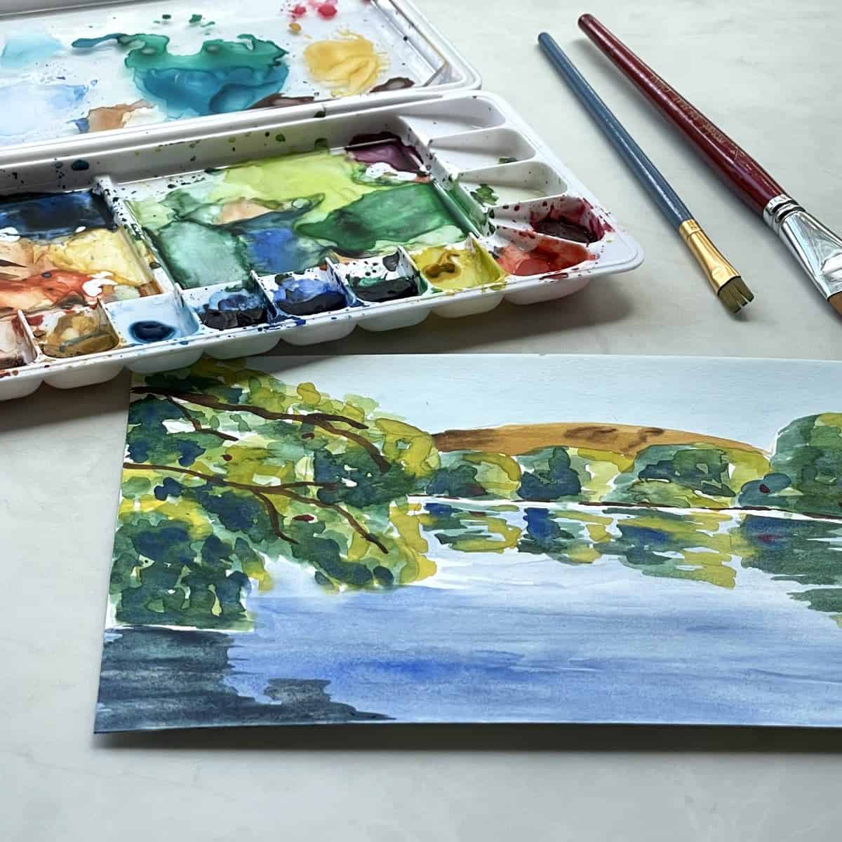 Painting of a watercolor lake scene with trees, a hill and reflections on the water next to paint brushes and a watercolor palette.