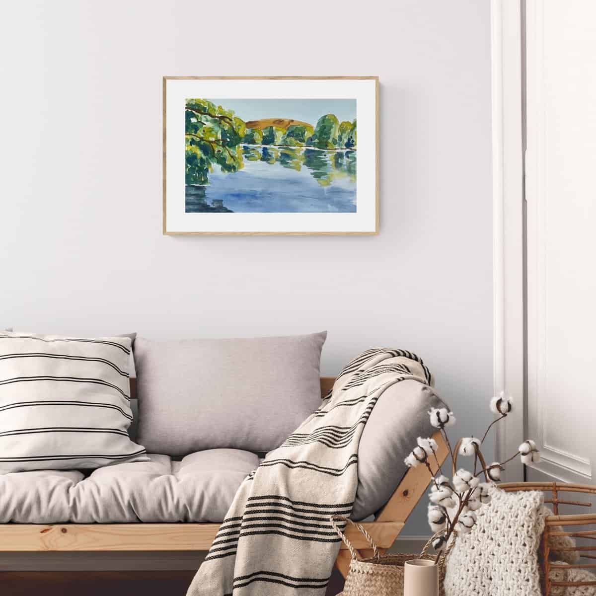 Painting of a watercolor lake scene with trees, a hill and reflections on the water hanging on a wall above a couch.