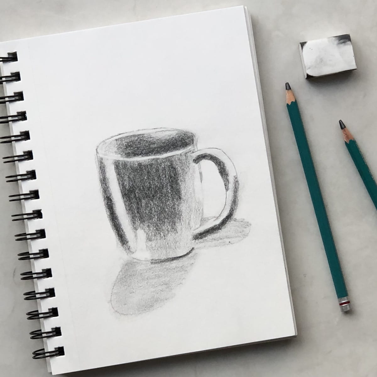Pencil sketch of a coffee mug on a spiral-bound sketchbook with drawing pencils and an eraser.