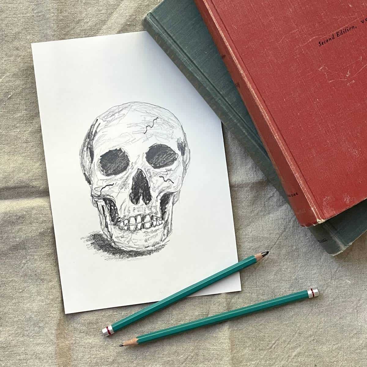 Skull drawing next to books and drawing pencils on a beige table cloth.