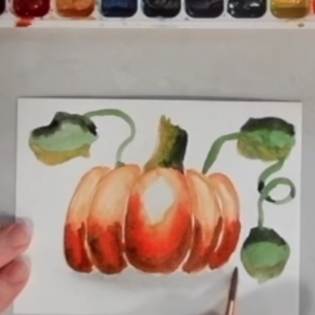 A hand painting a watercolor painting of a pumpkin.