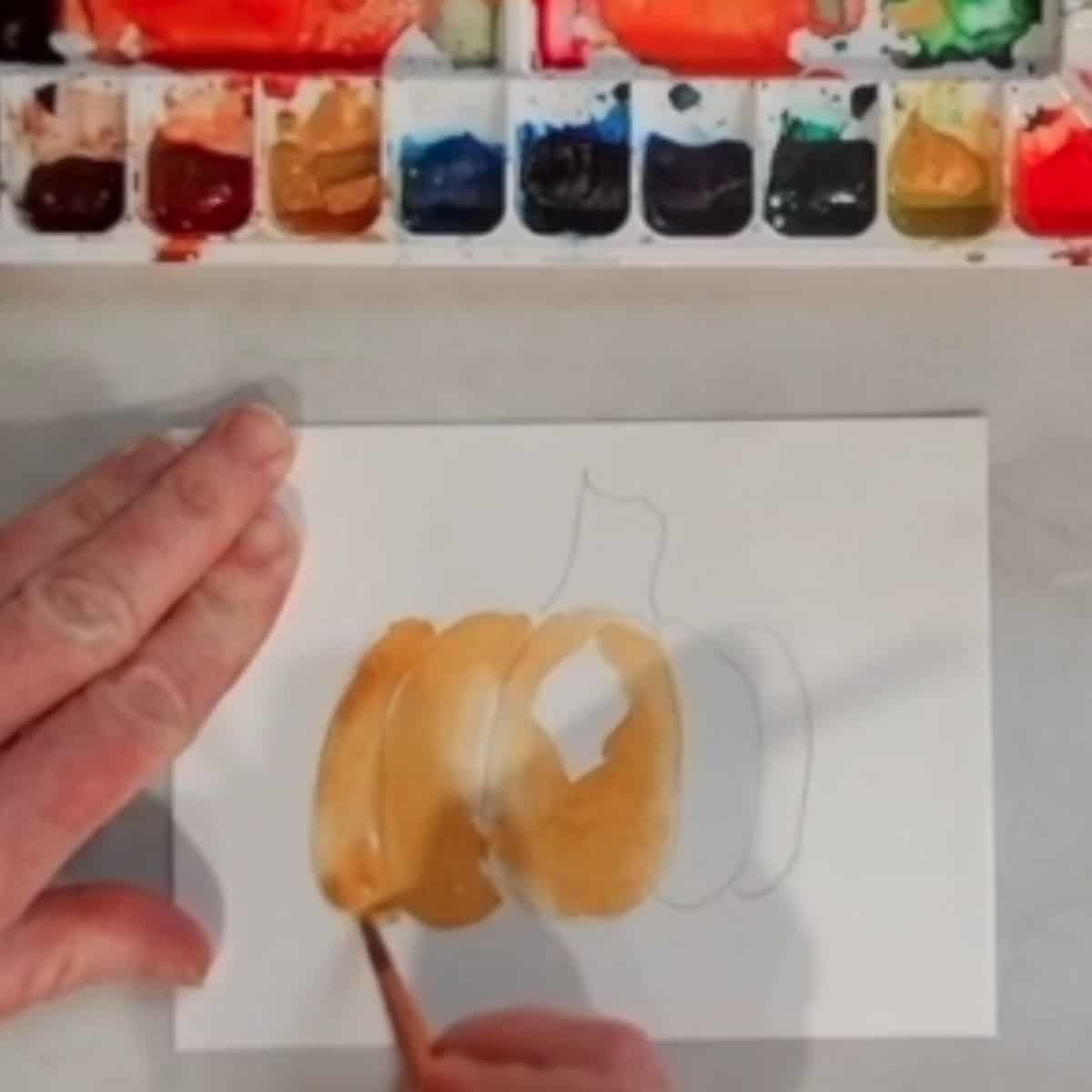 A hand painting a watercolor painting of a pumpkin.