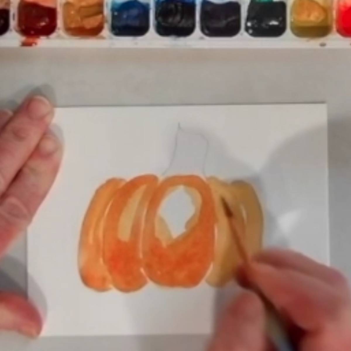 A hand painting a watercolor painting of a pumpkin.