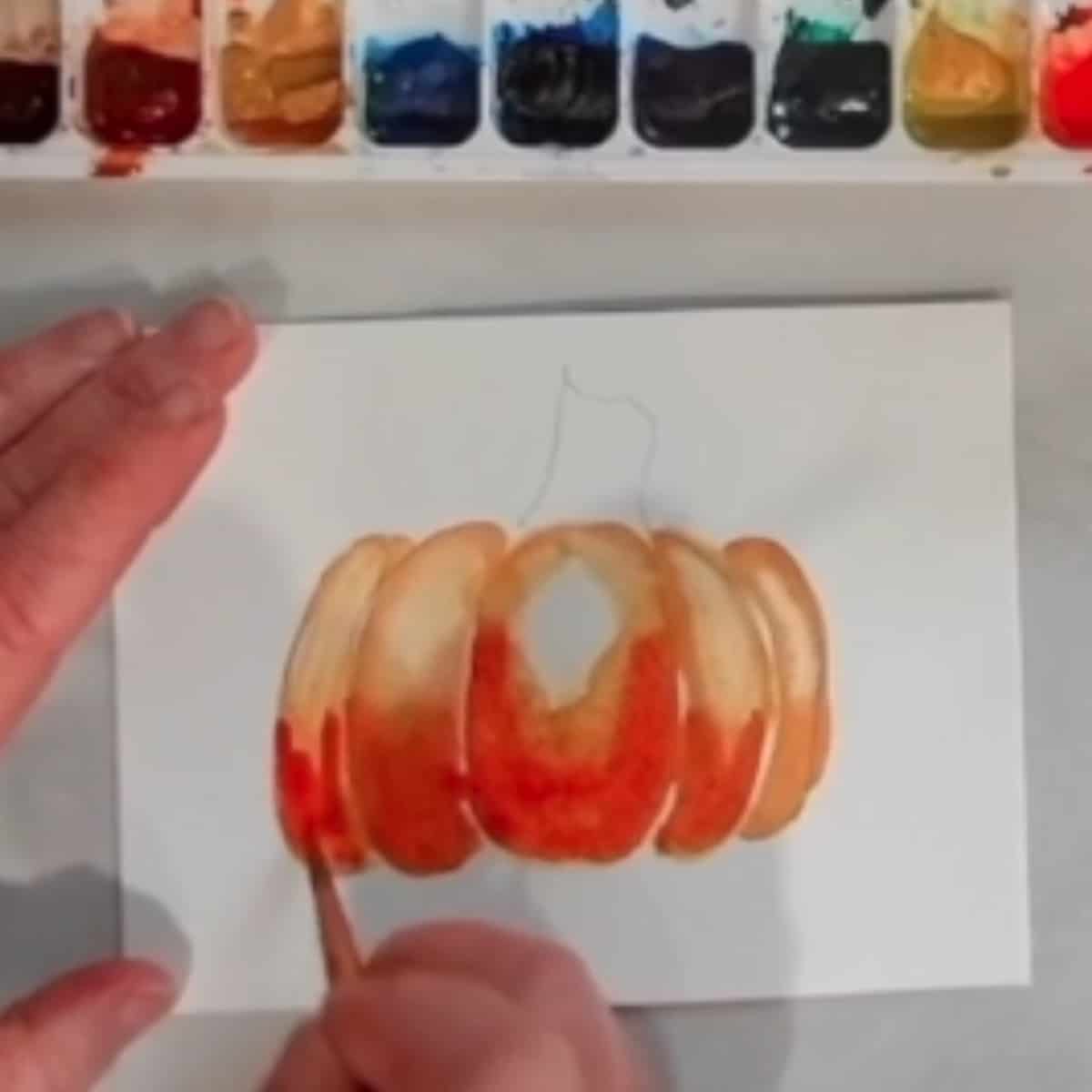 A hand painting a watercolor painting of a pumpkin.