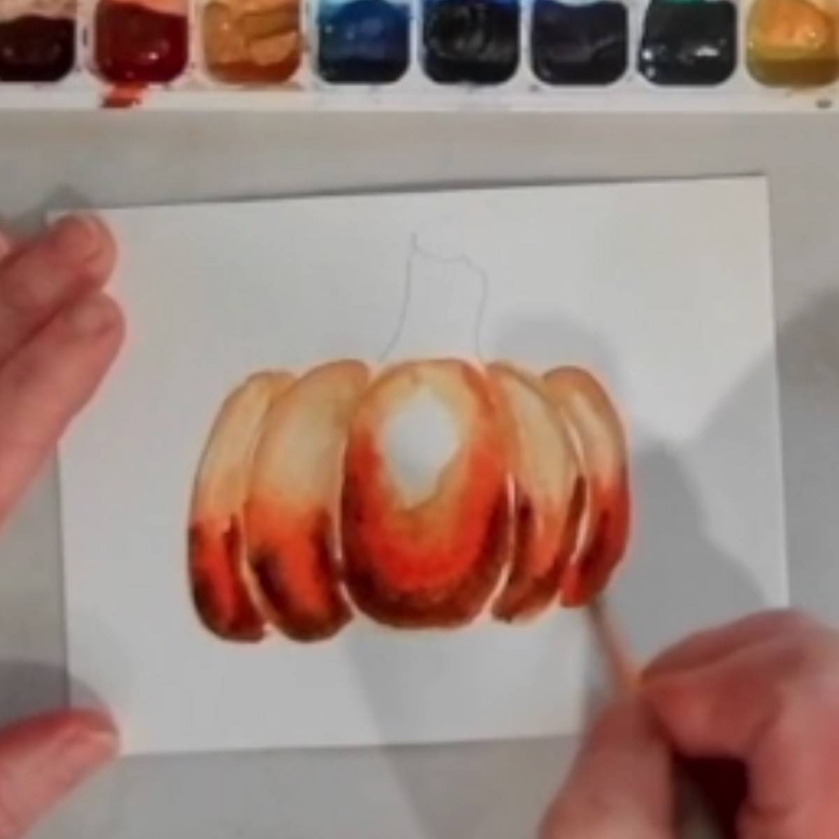 A hand painting a watercolor painting of a pumpkin.