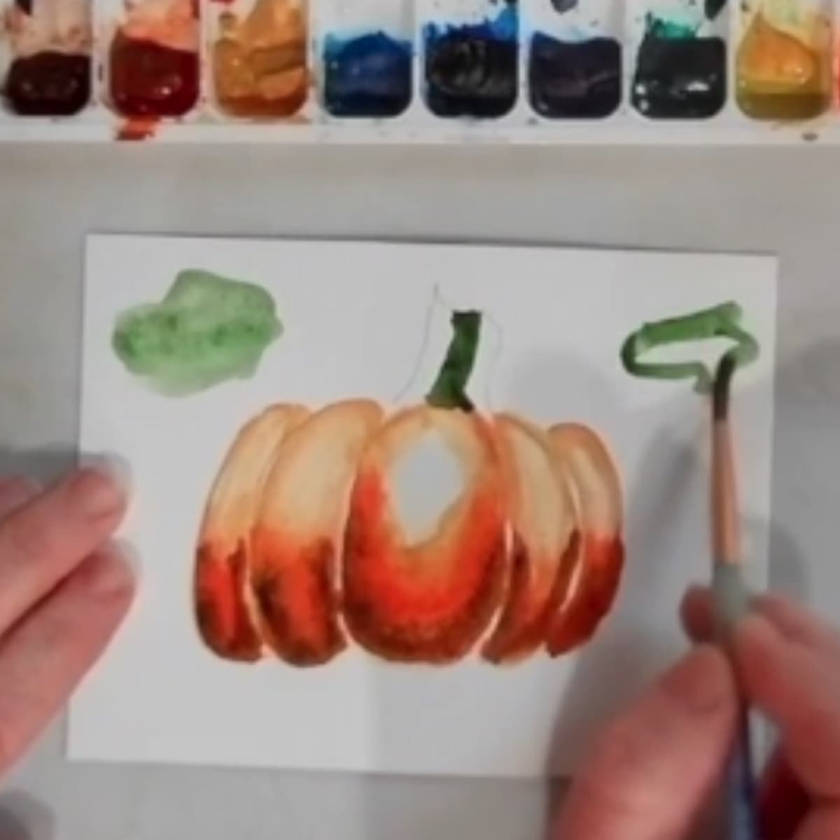 A hand painting a watercolor painting of a pumpkin.