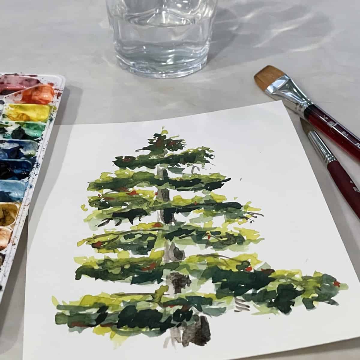 A watercolor painting of a pine tree next to paintbrushes, a cup of water, and a paint palette.