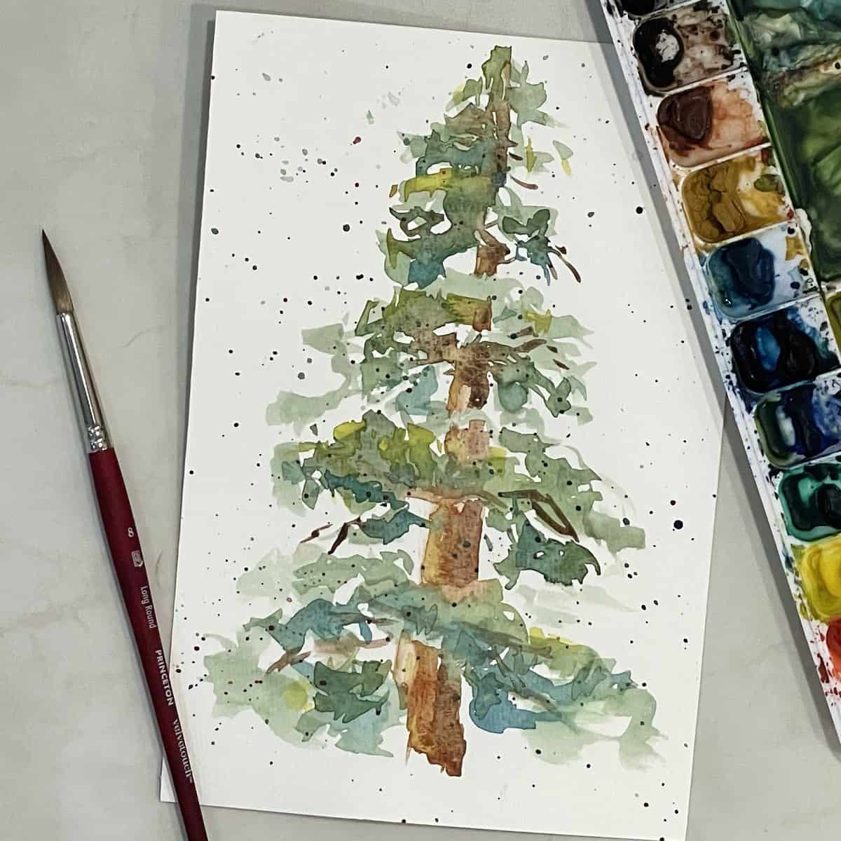 A watercolor painting of a pine tree with splattered paint on the painting, next to paintbrushes, a cup of water, and a paint palette.