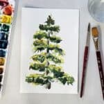 A watercolor painting of a pine tree next to paintbrushes, a cup of water, and a paint palette.