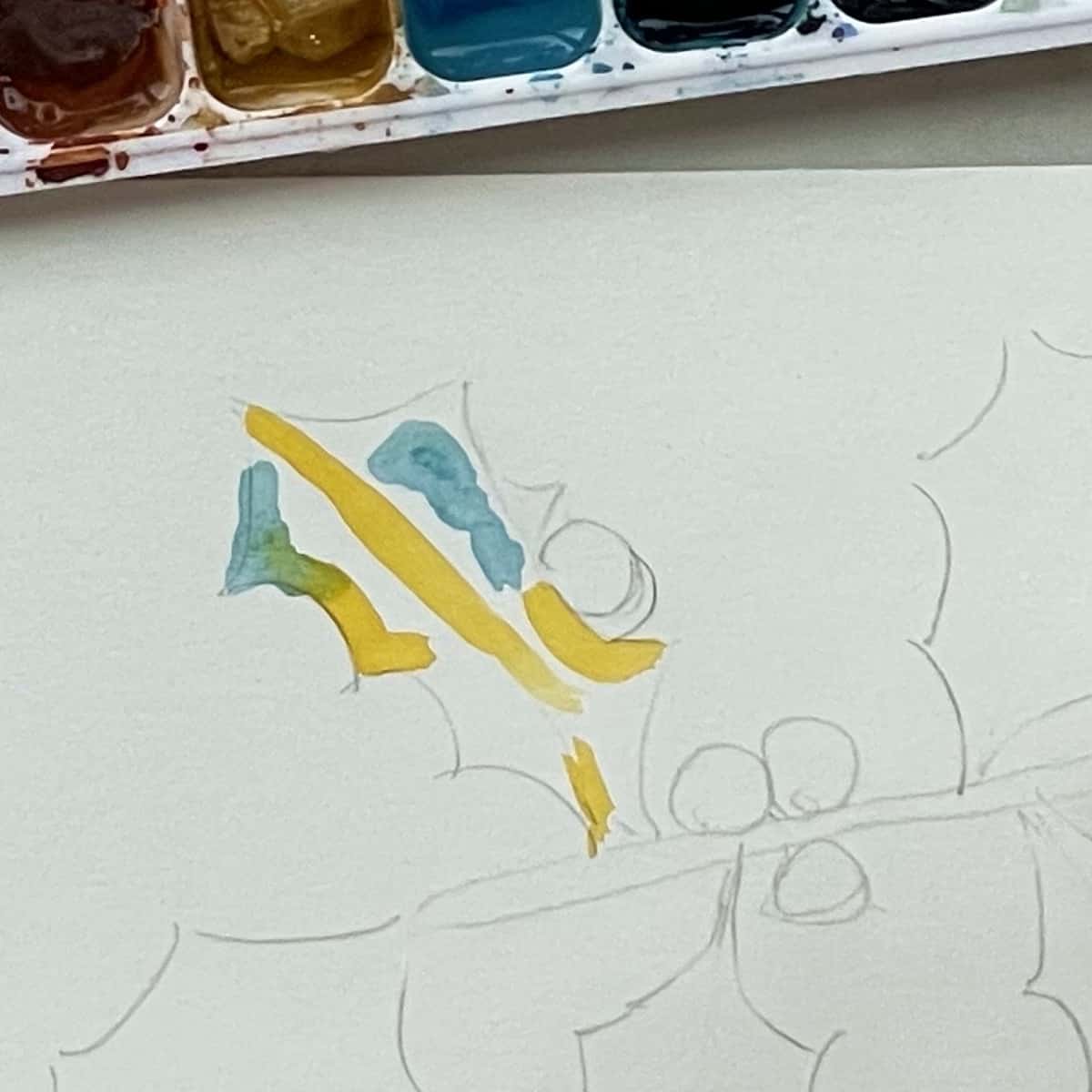 A holly leaf with some blue and yellow watercolor paint next to a paint palette. 