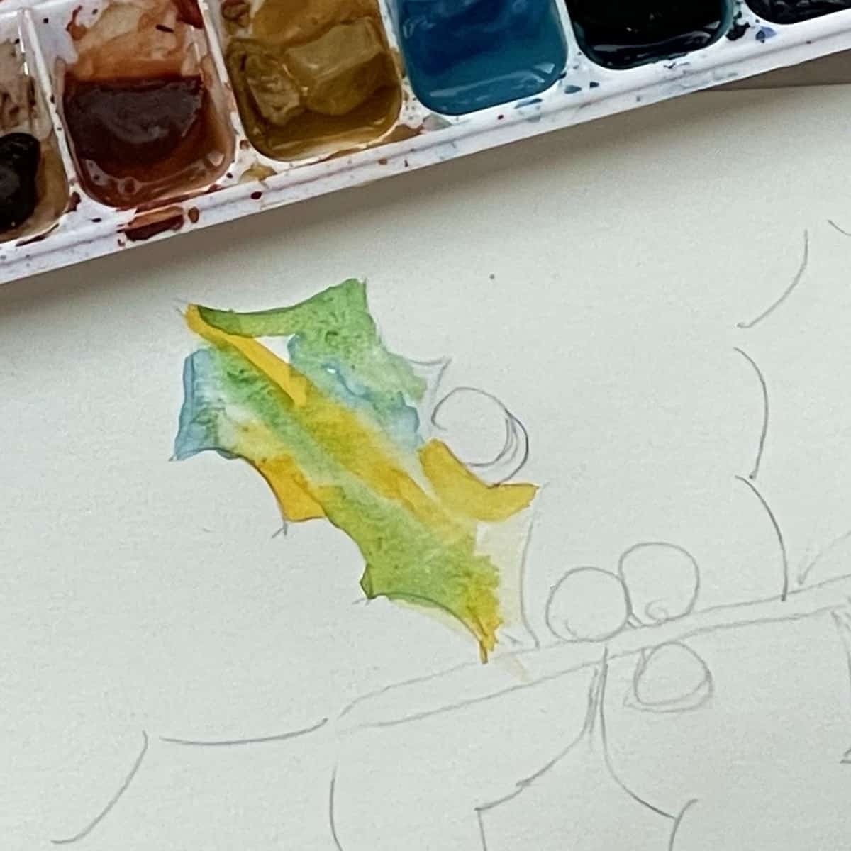 A holly leaf with some blue, yellow and green watercolor paint next to a paint palette. 