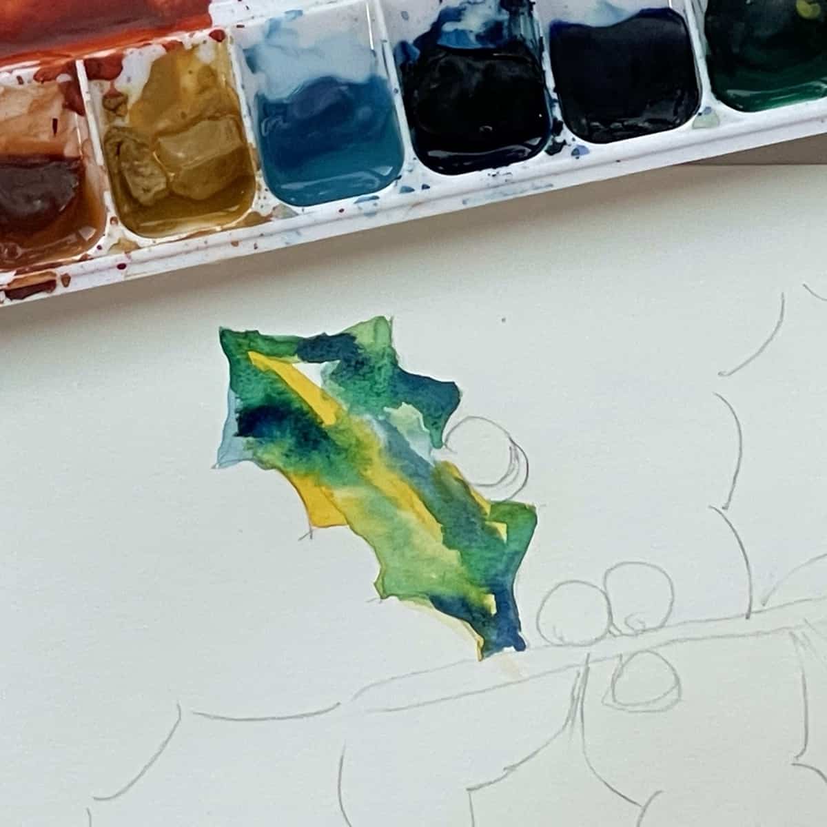 A holly leaf with some blue, yellow, green and dark blue watercolor paint next to a paint palette. 