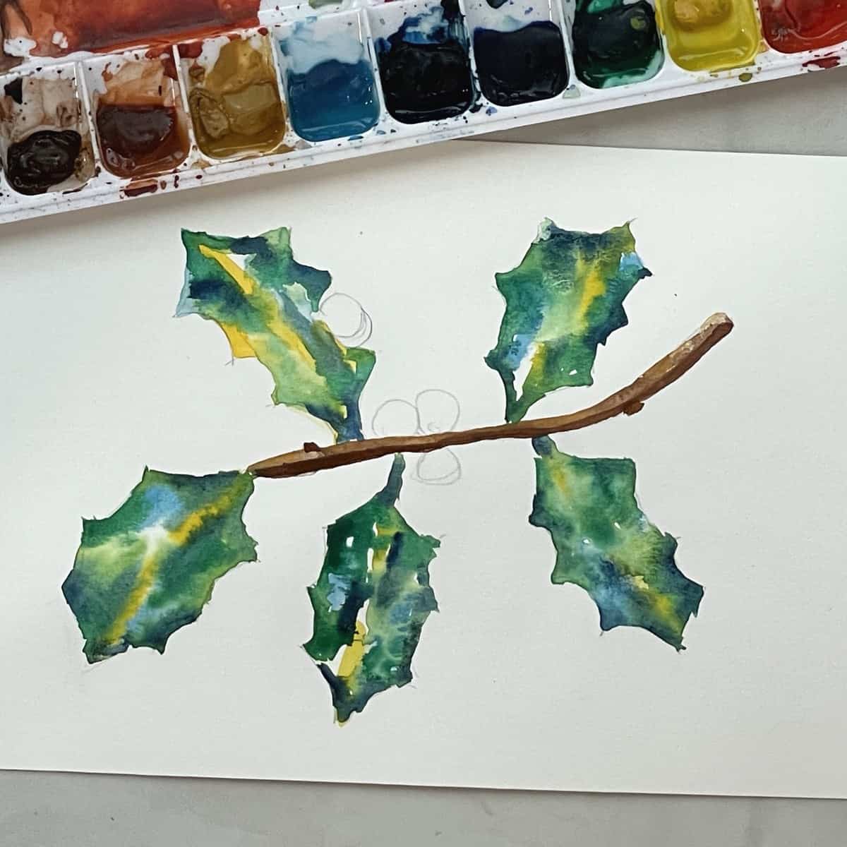 A watercolor painting of a sprig of holly with the leaves and stem painted in next to a watercolor palette.