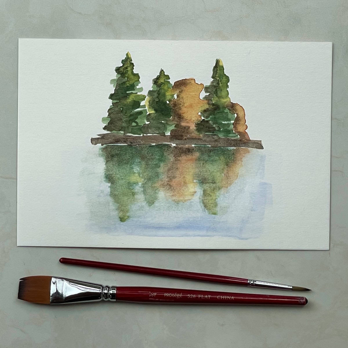 A painting of trees reflected on water in watercolor paint next to some paint brushes.