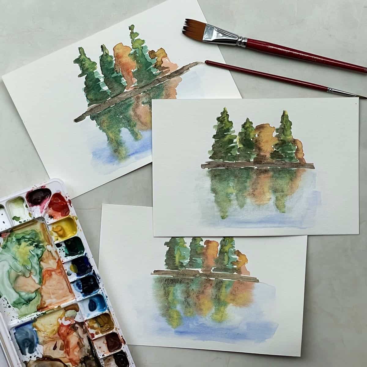 Three small watercolor paintings of reflections of trees on water next to paintbrushes and a watercolor paint set.