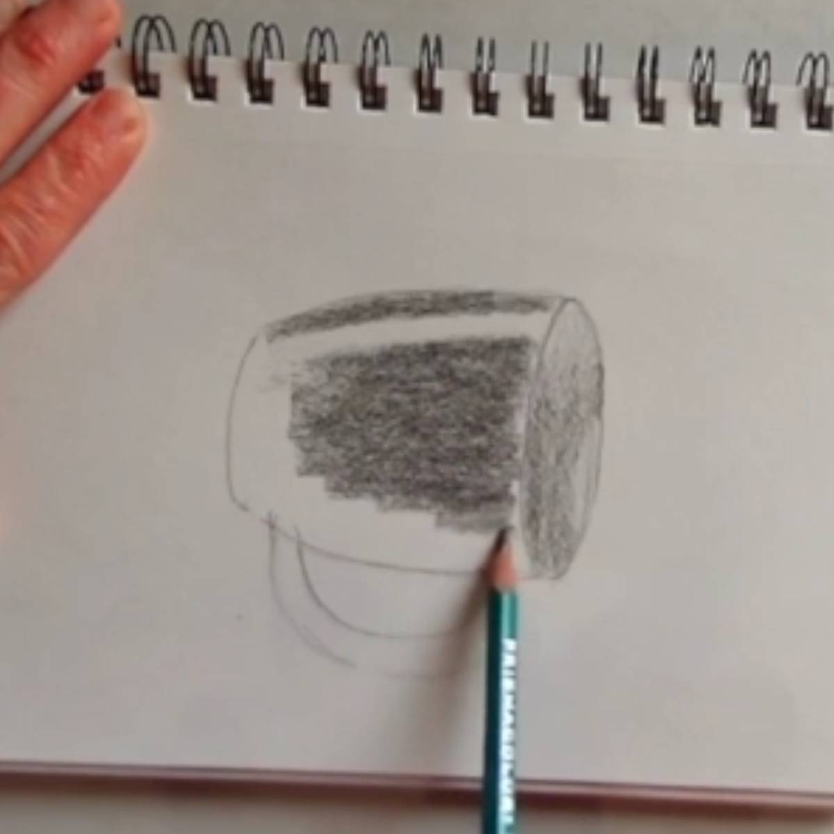 Shading in a sketch of a coffee mug in pencil with the drawing turned on the side for easier pencil movement.