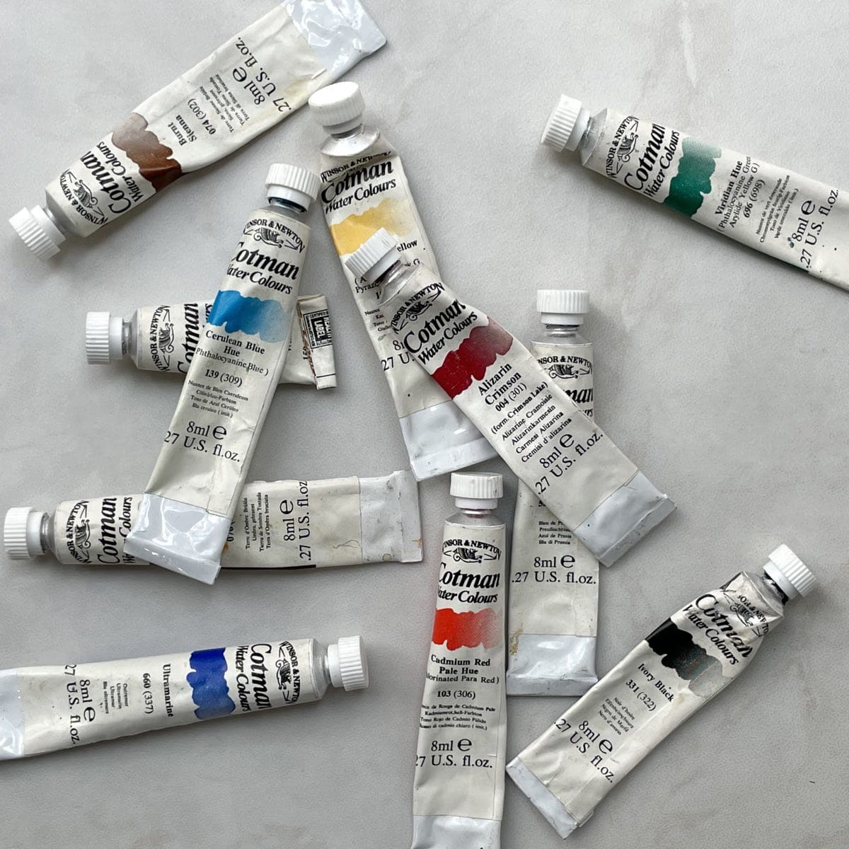 Eleven watercolor paint tubes scattered on a white table top.