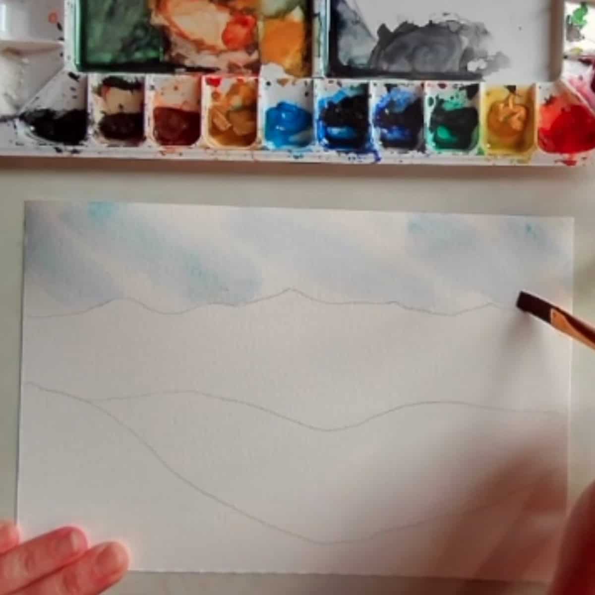 Artist's hand paints in the sky of a mountain landscape with a watercolor paint palette. 