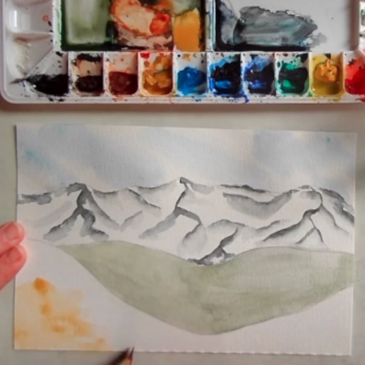 An artist working on painting a mountain landscape next to a watercolor paint palette. 