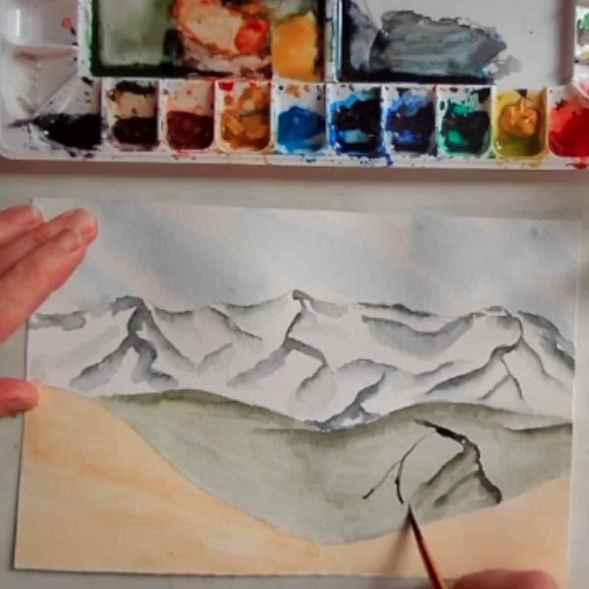 An artist painting a green mountain in a landscape painting next to a watercolor paint palette. 