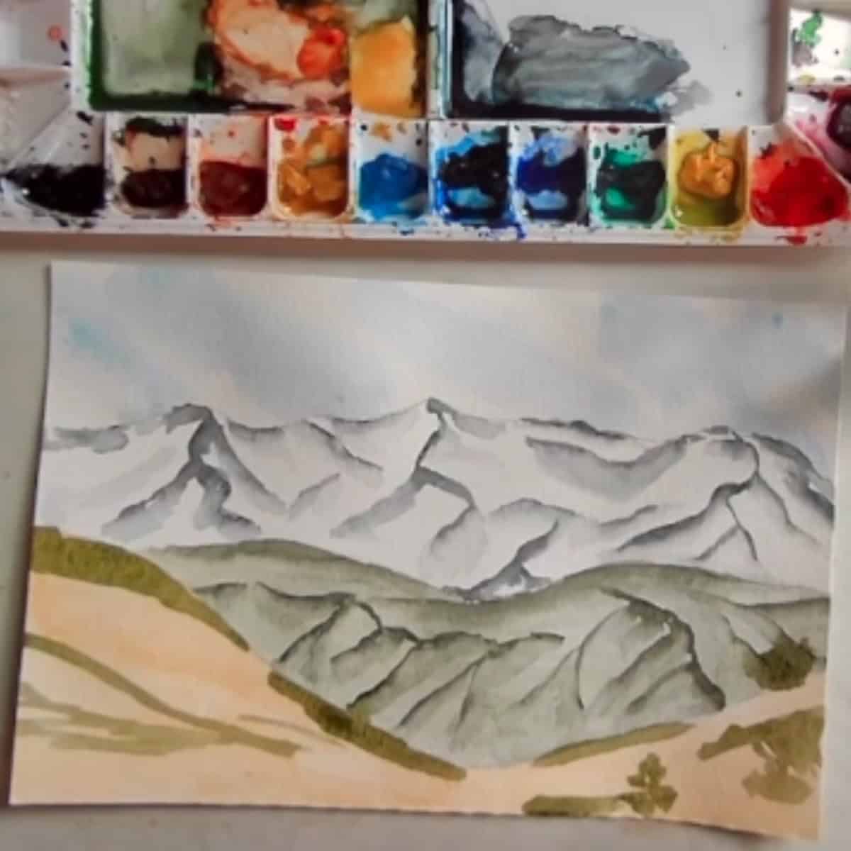 An almost complete painting of a mountain landscape with green details in the foreground next to a watercolor paint palette. 