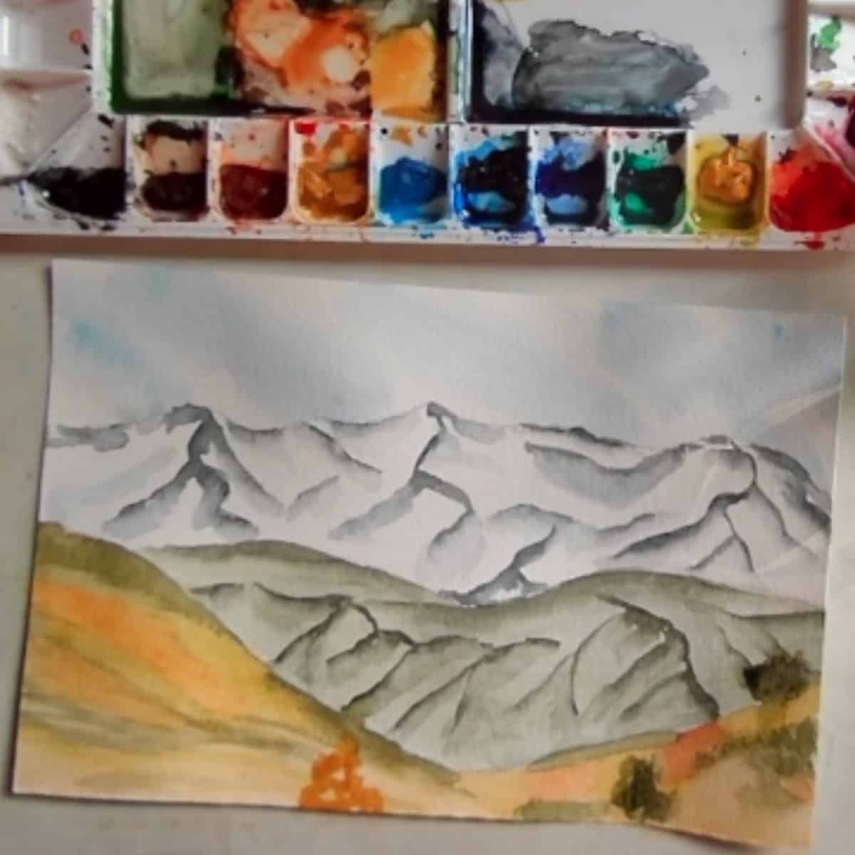 A completde painting of a mountain landscape with next to a watercolor paint palette. 