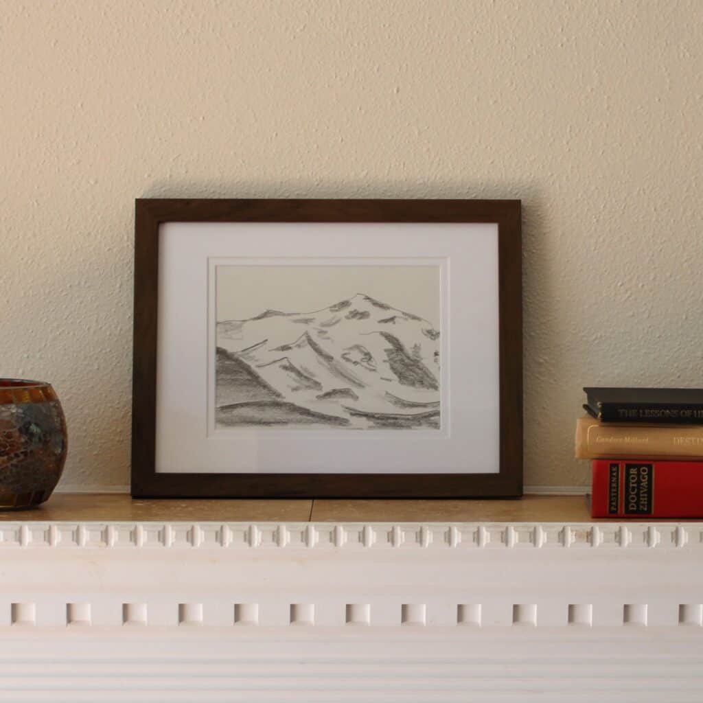 Mountain landscape framed on a mantel piece next to books and vase