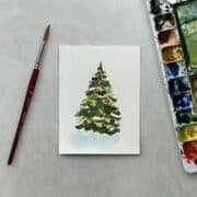 A watercolor painting of a Christmas tree next to a paintbrush and palette.