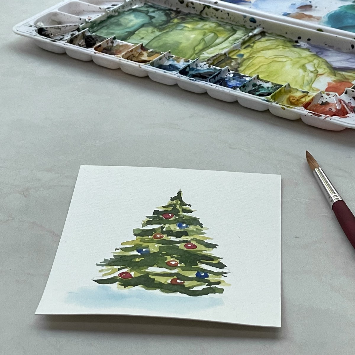 A watercolor painting of a Christmas tree at a foreshortened angle next to a paintbrush and palette.