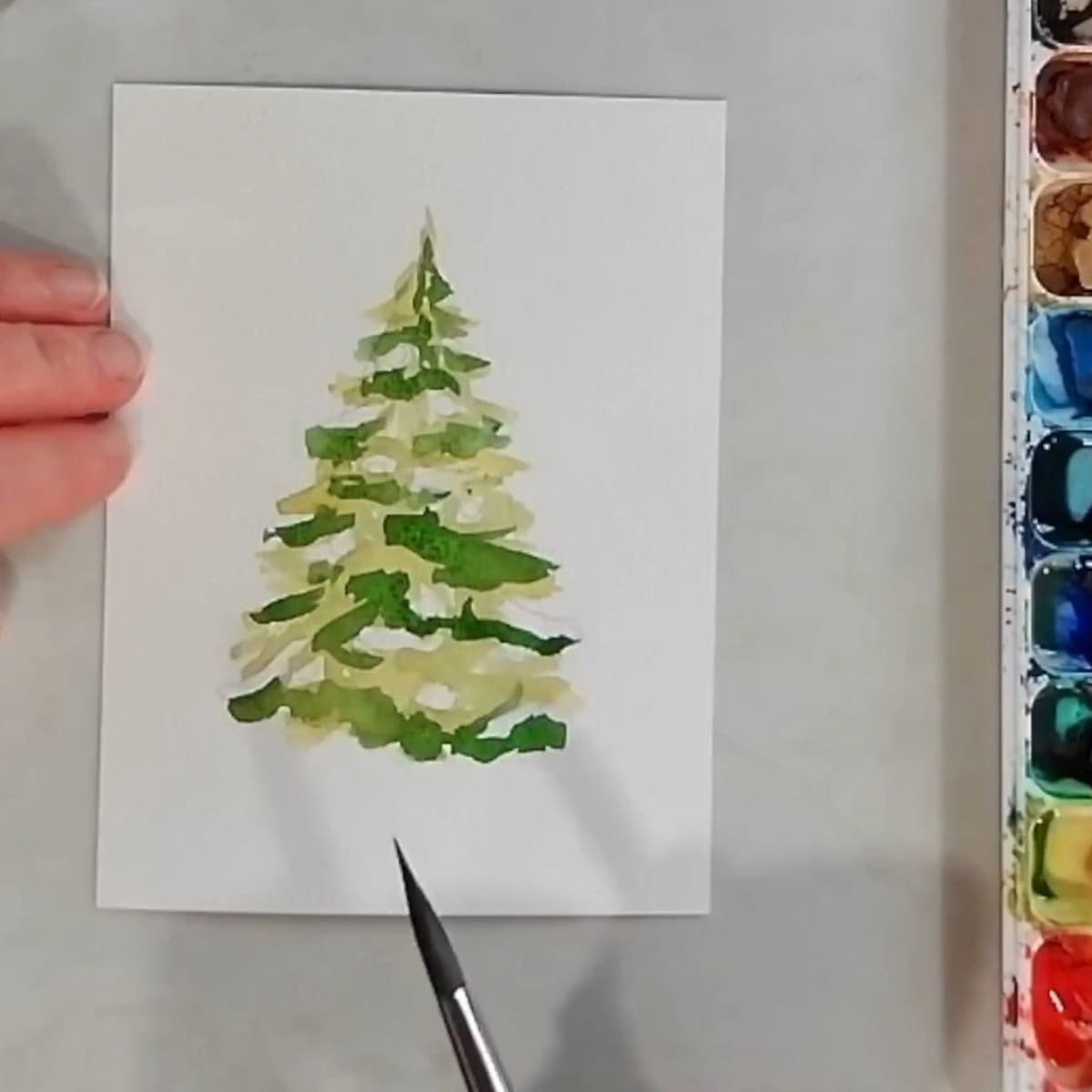 A Christmas tree painted with two layers of watercolor paint, light green and medium green, next to a paint brush and palette.