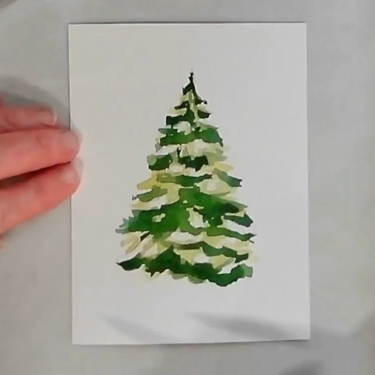 A watercolor painting of a basic Christmas tree with an artist's hand.