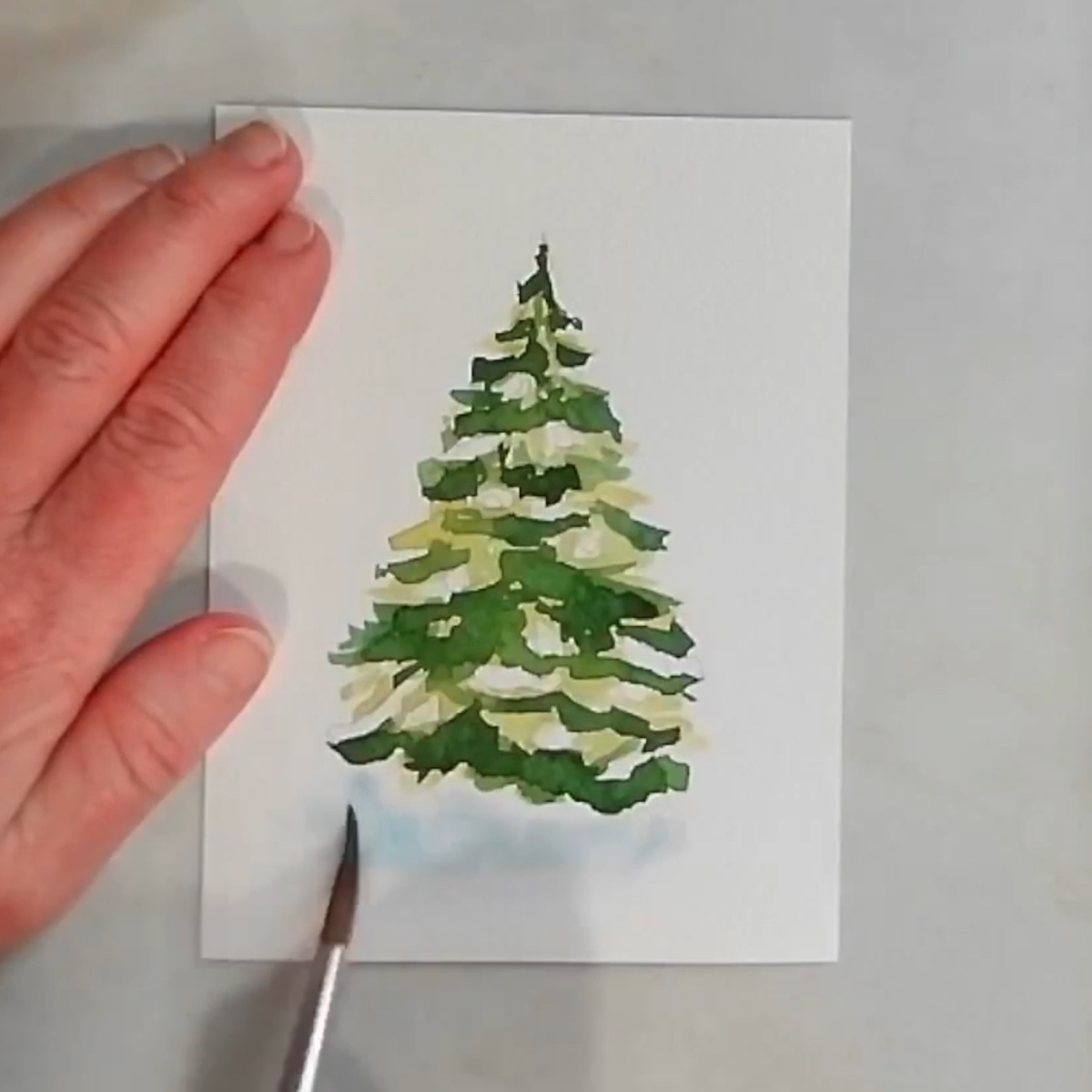 A watercolor painting of a basic Christmas tree with an artist's hand adding a light blue shadow in the foreground.