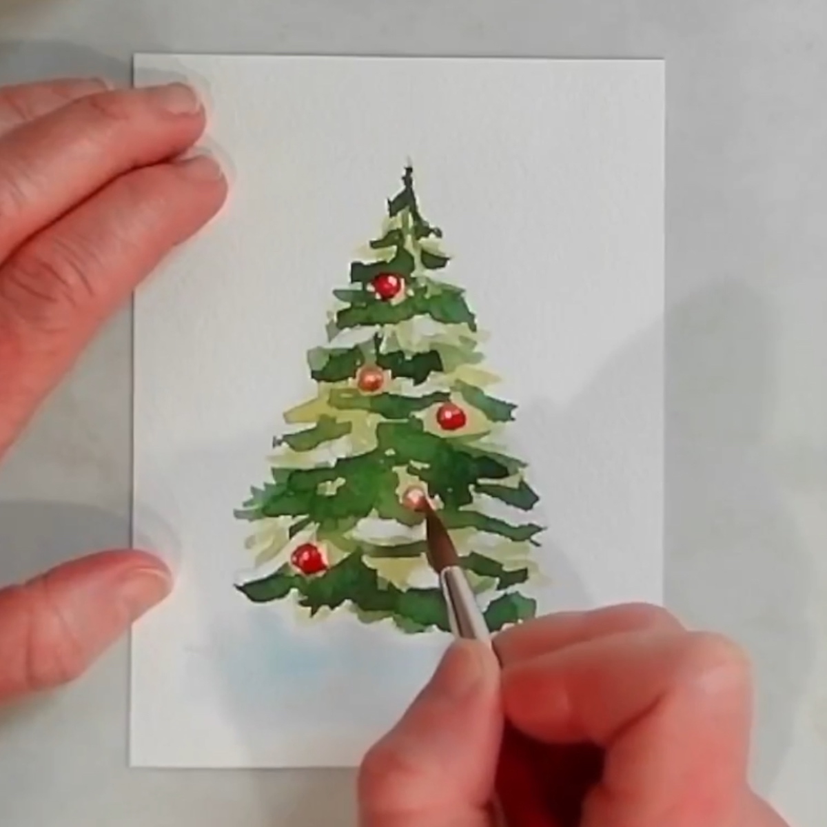 An artist's hand painting colorful ornaments on a Christmas tree in watercolor.