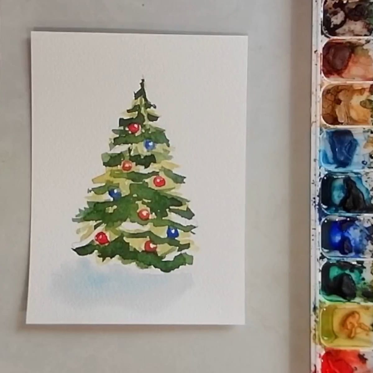 A completed watercolor painting of a Christmas tree with colorful ornaments  and patches of snow next to a paint palette. 