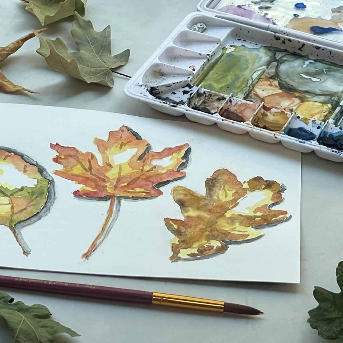 A watercolor painting of three colorful fall leaves next to a paintbrush, paint palette, and leaves.