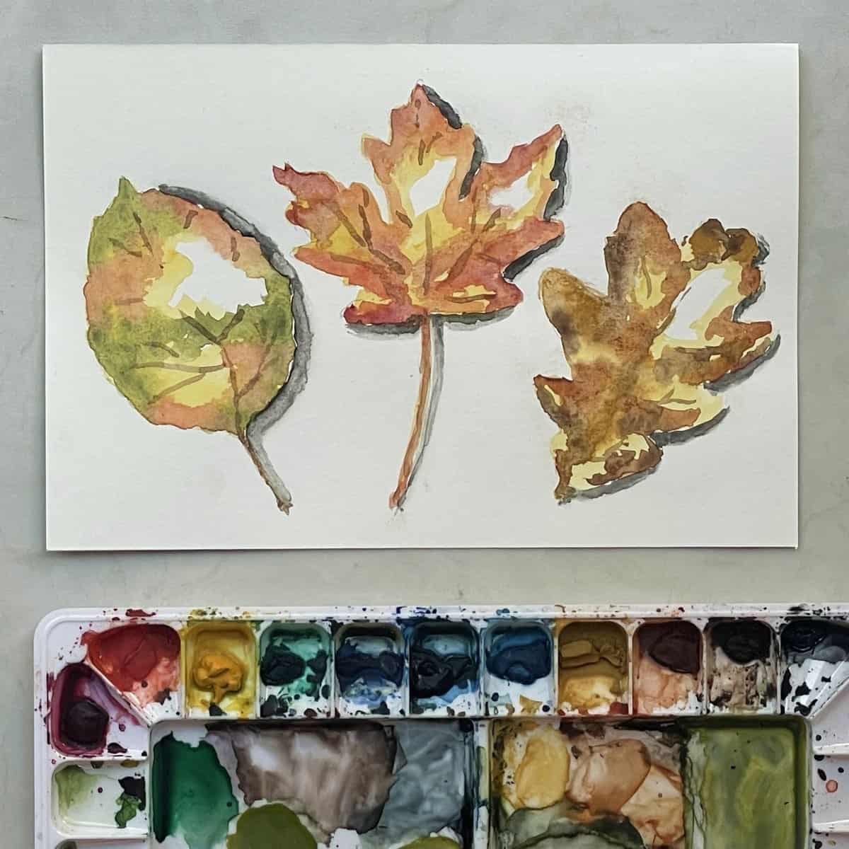 A watercolor painting of three colorful autumn leaves next to a paint palette.