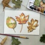 A watercolor painting of three colorful fall leaves next to a paintbrush, paint palette, and leaves.